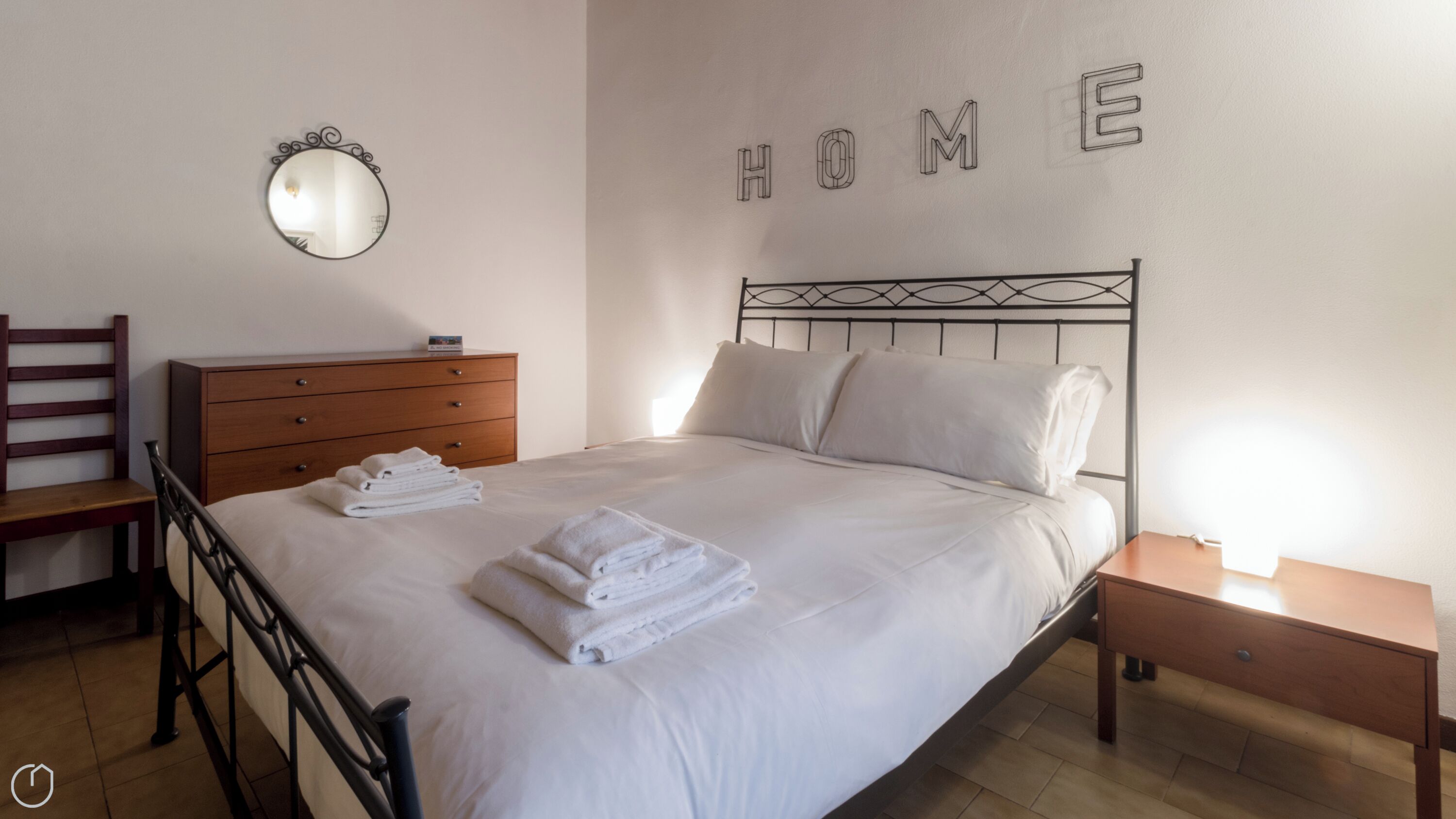 A neatly made bed with folded towels, a wooden dresser, a bedside table with a lamp, and a wall-mounted mirror.