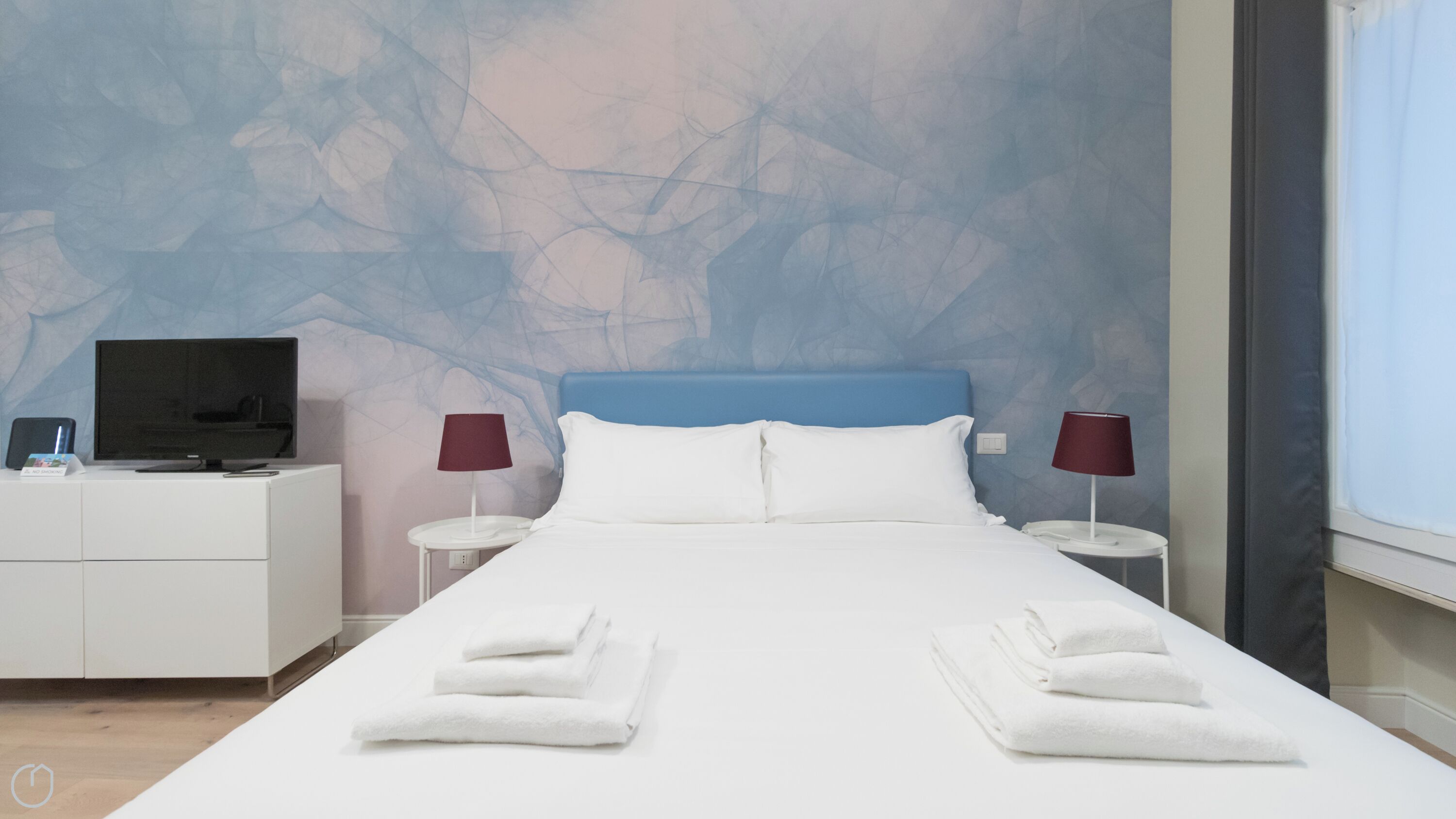 A hotel room with a bed, two bedside tables, a TV, and a wall with a tree pattern.