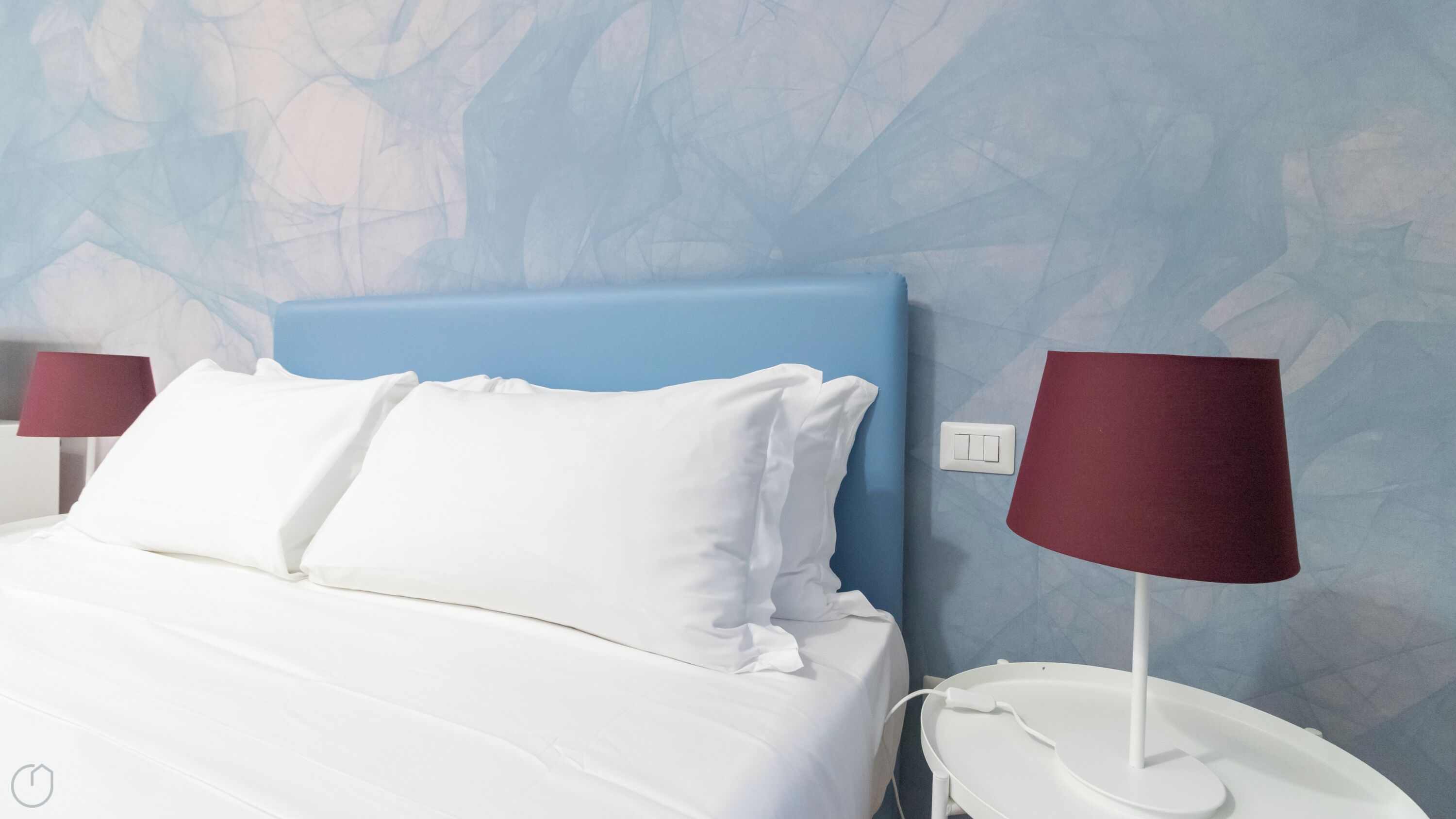 A neatly made bed with white linens and a blue headboard, a white bedside table with a red lamp, and a wall with a textured pattern.
