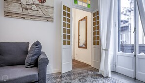 A room with a grey sofa, a patterned rug, a large mirror, and a view of the street through a window.