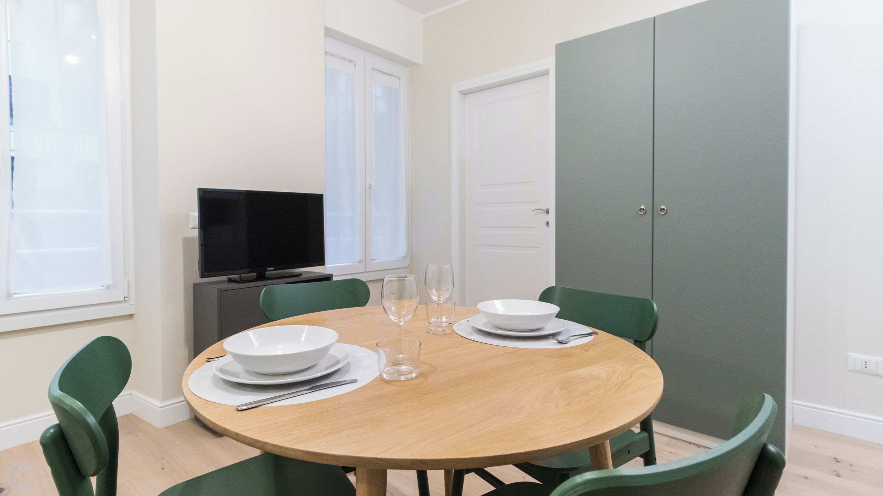 A dining area with a round wooden table, green chairs, white plates, and a television on a cabinet.