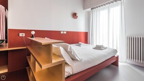 A modern hotel room with a wooden desk, a bed with white linens, a wall-mounted light, and a window with sheer curtains. - Italianway - Bassini 31 (Milan)