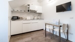 Een moderne keuken met witte kastjes, een magnetron, waterkoker en koffiezetapparaat. Er staat een eettafel met stoelen en een flatscreen-tv hangt aan de muur.