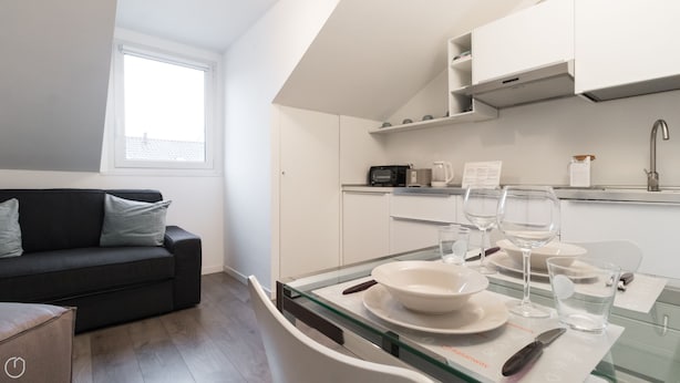 A modern kitchen with white cabinets, a microwave, and a coffee maker. There is a dining table with plates, glasses, and cutlery. A black sofa is against the wall with a window above it. - Italianway - Corso Como 9 (Milan)