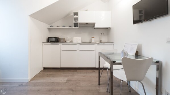 Een compacte keuken met witte kastjes, een magnetron, een waterkoker en een kleine eettafel met stoelen.