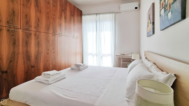 Habitación de hotel con un armario de madera, una cama bien hecha con sábanas blancas, una mesita de noche con lámpara y una ventana con cortinas de gasa.