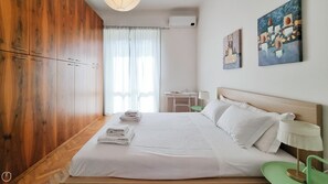 Quarto com armário de madeira, cama com roupa de cama branca, escrivaninha com cadeira e obras de arte na parede.
