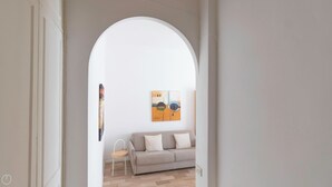 Un pasillo con un arco blanco que da a una sala de estar con un sofá color beige y un cuadro en la pared.