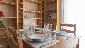 Una mesa de comedor con platos, vasos y cubiertos, rodeada de sillas de madera y estantes con utensilios de cocina.