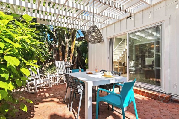 Eine Außenpatio mit einem weißen Tisch und türkisfarbenen Stühlen, umgeben von einer weißen Pergola und üppiger Bepflanzung.