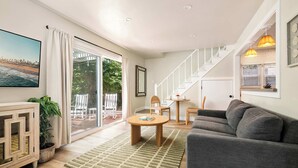 Un salon moderne avec un canapé gris, une table basse en bois et un tapis à motifs verts. Un escalier avec des rampes blanches mène à un espace extérieur doté d’une porte en verre.