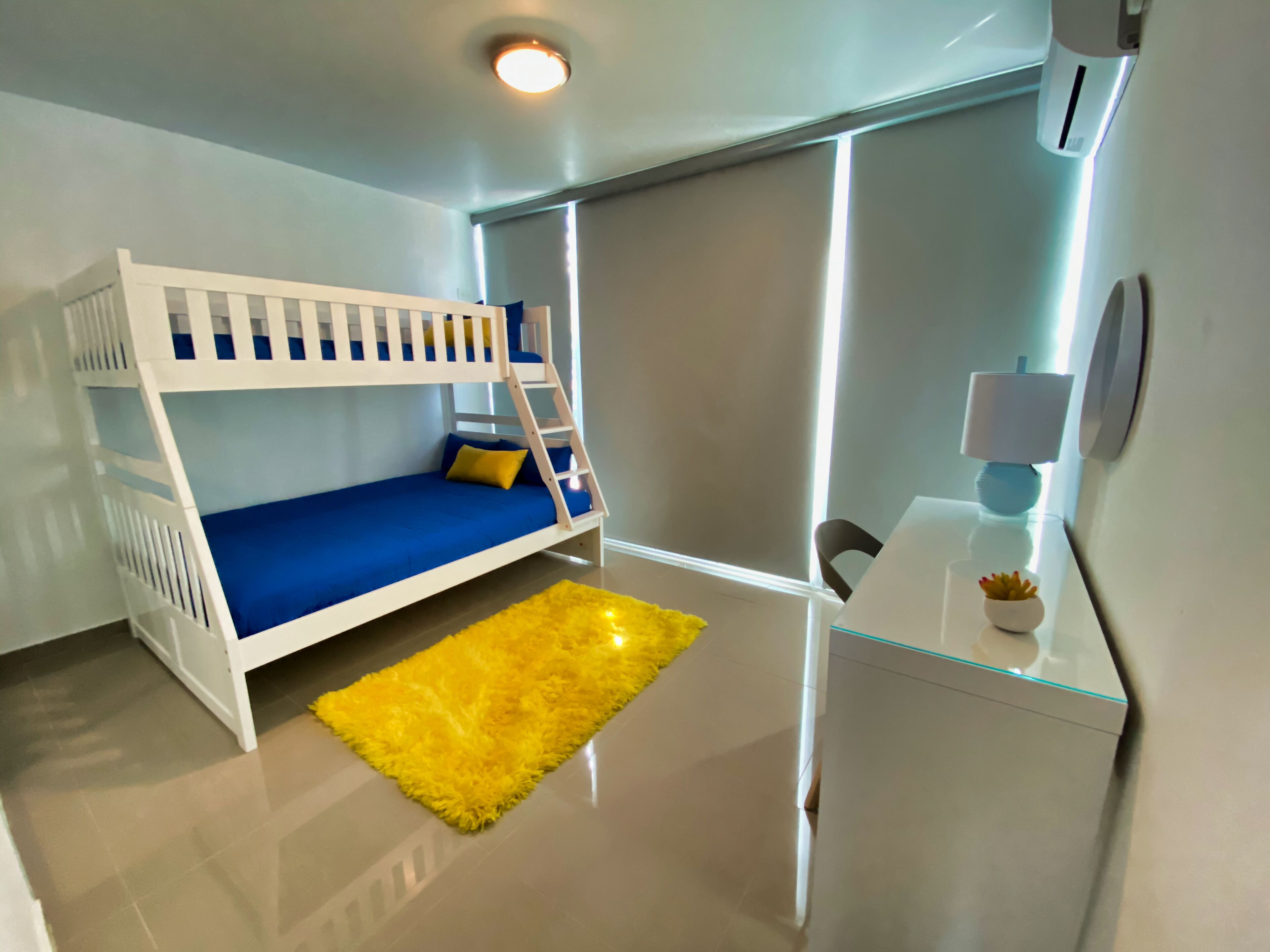 Una cama individual superpuesta con colchón azul, alfombra amarilla, escritorio con lámpara y espejo.