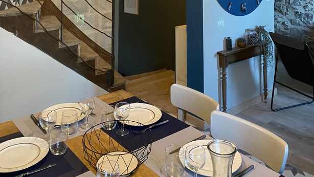 Een eethoek met een houten tafel gedekt voor vier personen, een grote wandklok en een trap met een geometrisch patroon op de muur.