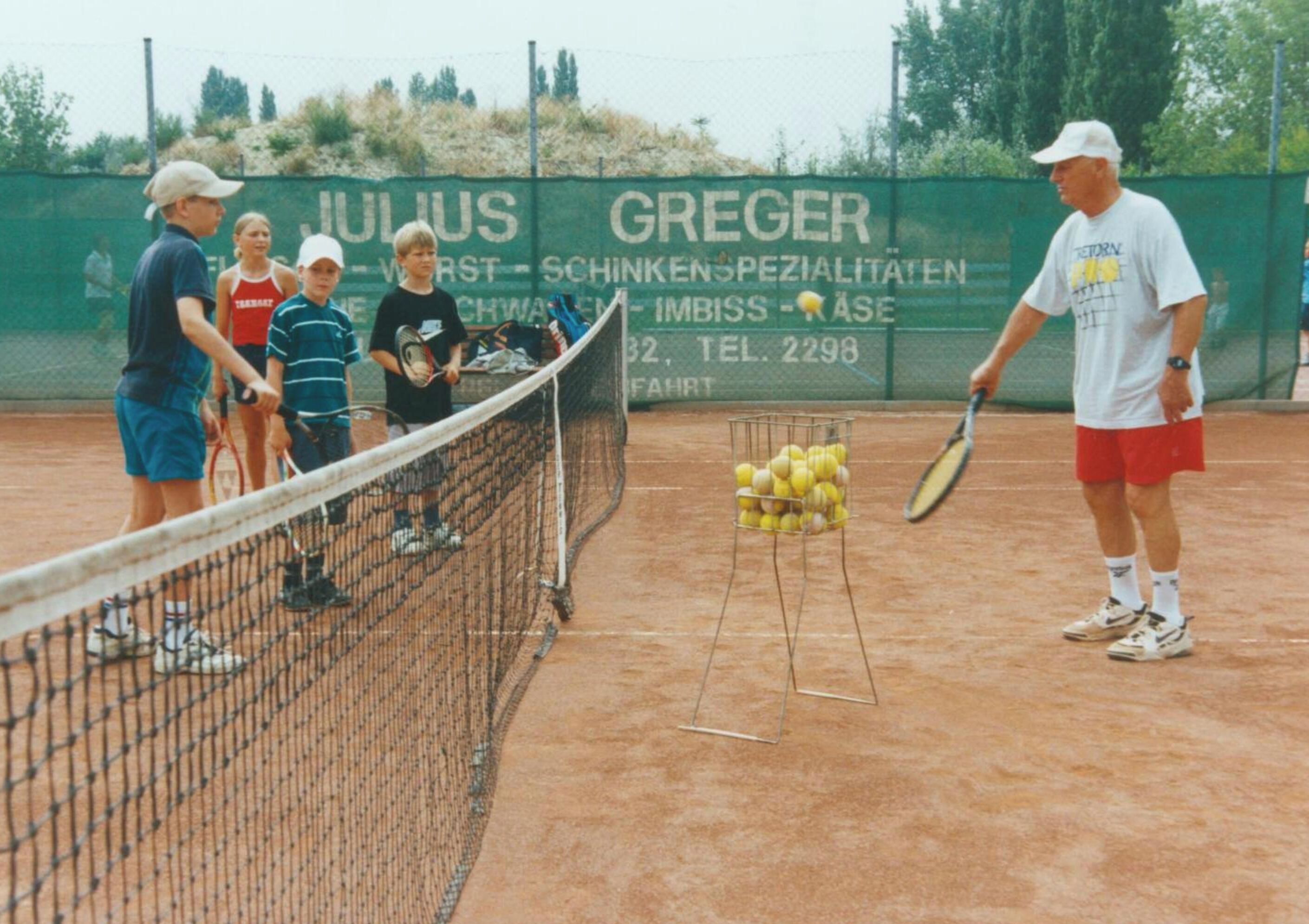 A tennis court with a net, a rack of tennis balls, and a person holding a tennis racket.