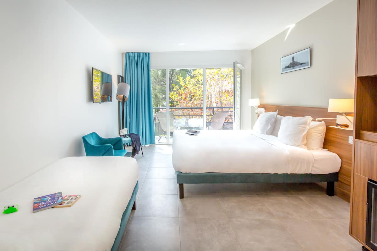 Habitación de hotel con dos camas, un balcón con vistas a árboles, una silla y una mesita.