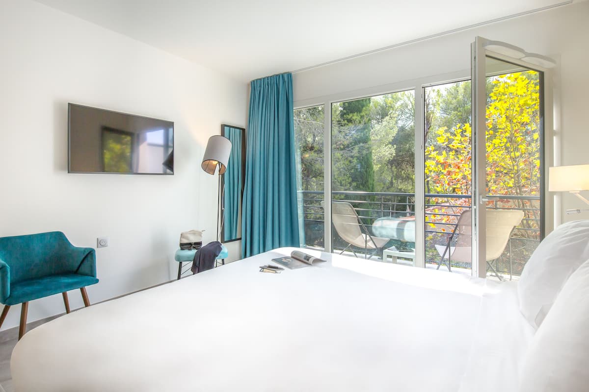 Una habitación de hotel moderna con una cama grande, un televisor montado en la pared, un balcón con sillas y vistas a árboles otoñales.