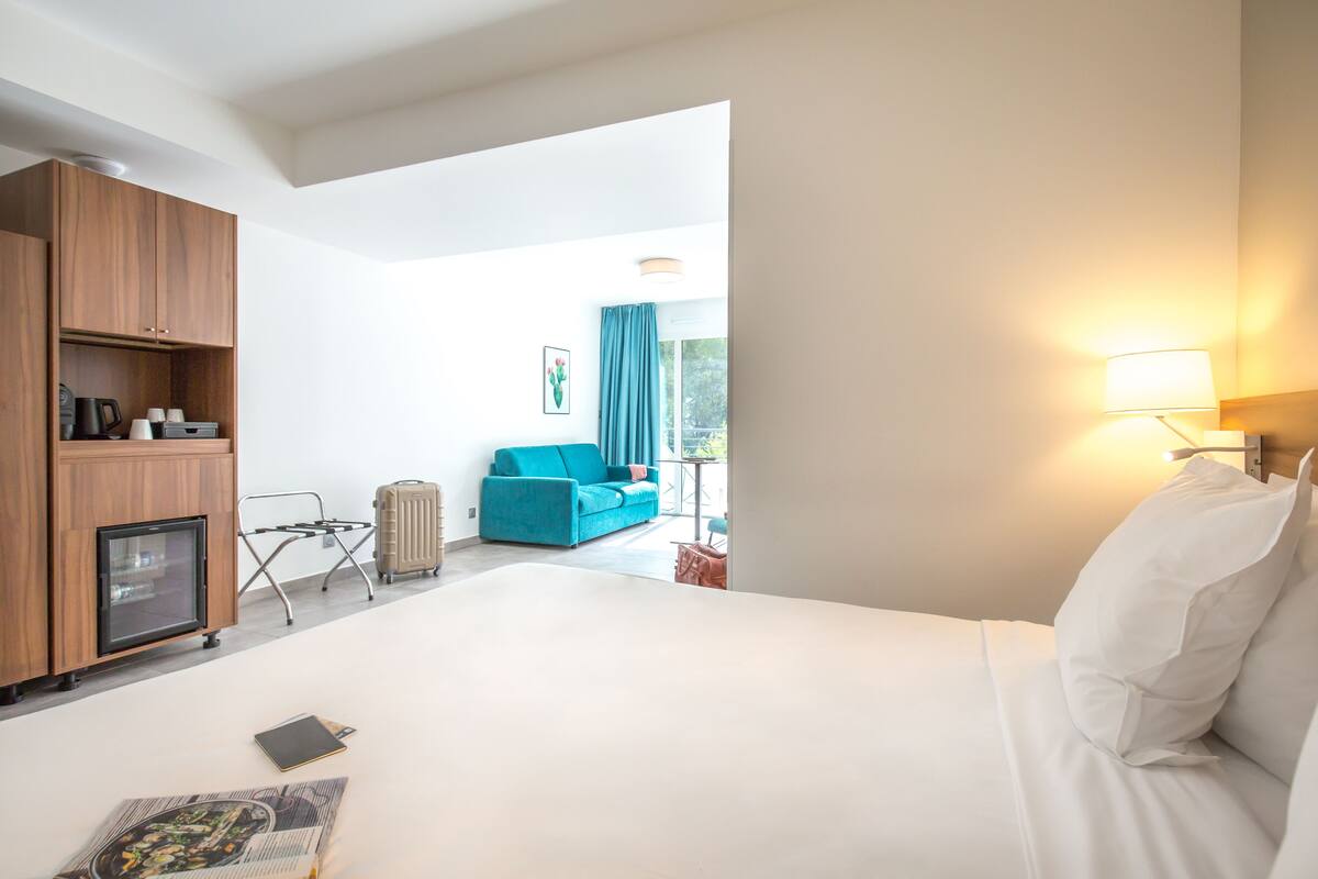 Habitación de hotel con cama, escritorio con lámpara, armario, y vista a un sofá azul y cortinas al fondo.