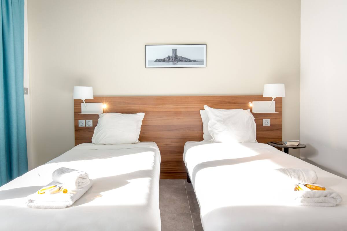 Habitación de hotel con dos camas, cada una con ropa de cama y almohadas blancas, cabeceros de madera, lámparas de noche, un cuadro en la pared y una mesita con un libro y una botella.