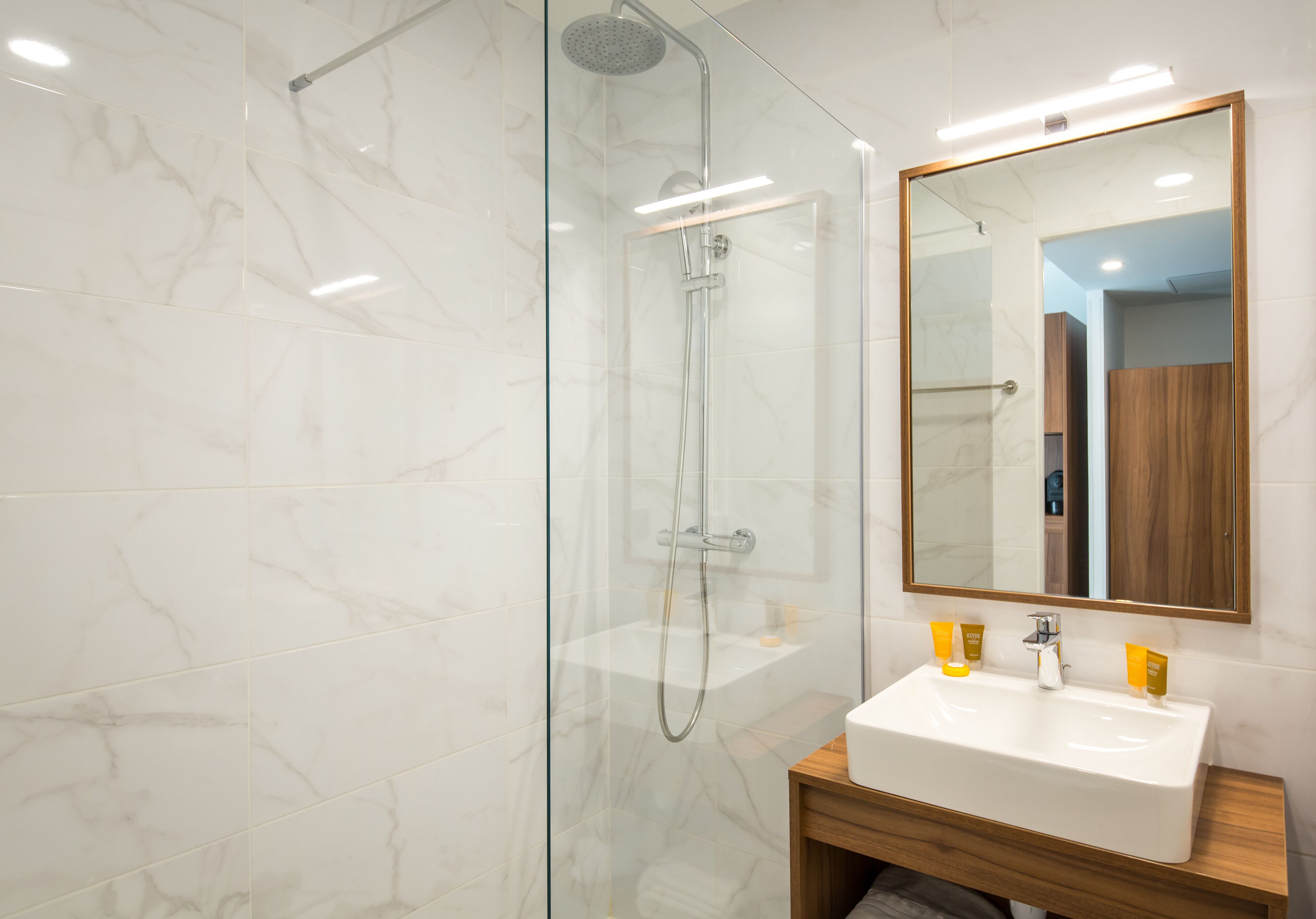 Un baño moderno con ducha envuelta en vidrio, tocador de madera con lavamanos blanco y un espejo grande.