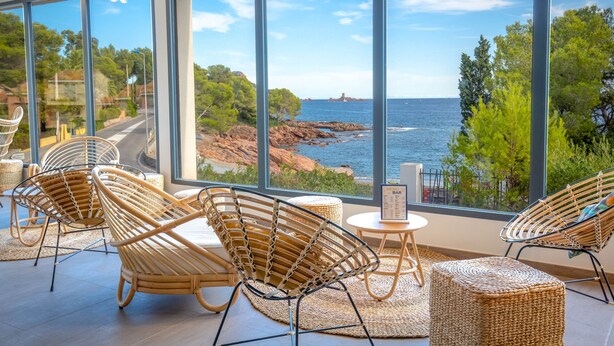 A room with large windows overlooking a coastal landscape, wicker furniture, and a small table with a menu. - SOWELL HÔTELS La Plage (Saint-Raphael)