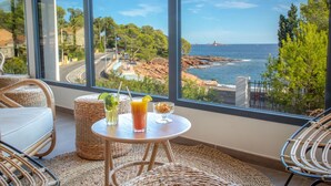 A balcony with wicker furniture, a table with drinks, and a view of the sea and rocky coastline. - SOWELL HÔTELS La Plage (Saint-Raphael)