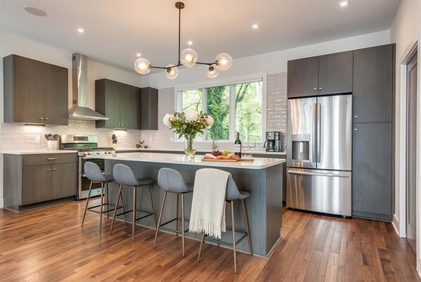 A modern kitchen with a central island, bar stools, a stainless steel refrigerator, and a large window providing natural light.