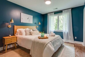 A bedroom with a bed, bedside lamps, a nightstand, a window with curtains, and a painting on the wall.
