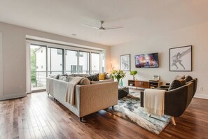 A spacious living room with hardwood floors, a large window, a ceiling fan, a sofa, a coffee table, and artwork on the walls.