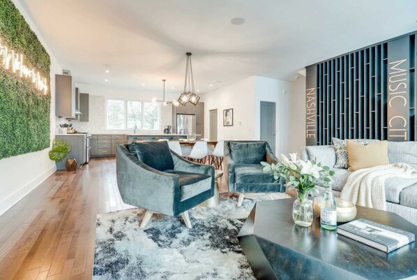 Una sala de estar moderna con un sofá, dos sillones, una mesa de centro y una alfombra. Se observa una cocina al fondo con un comedor.