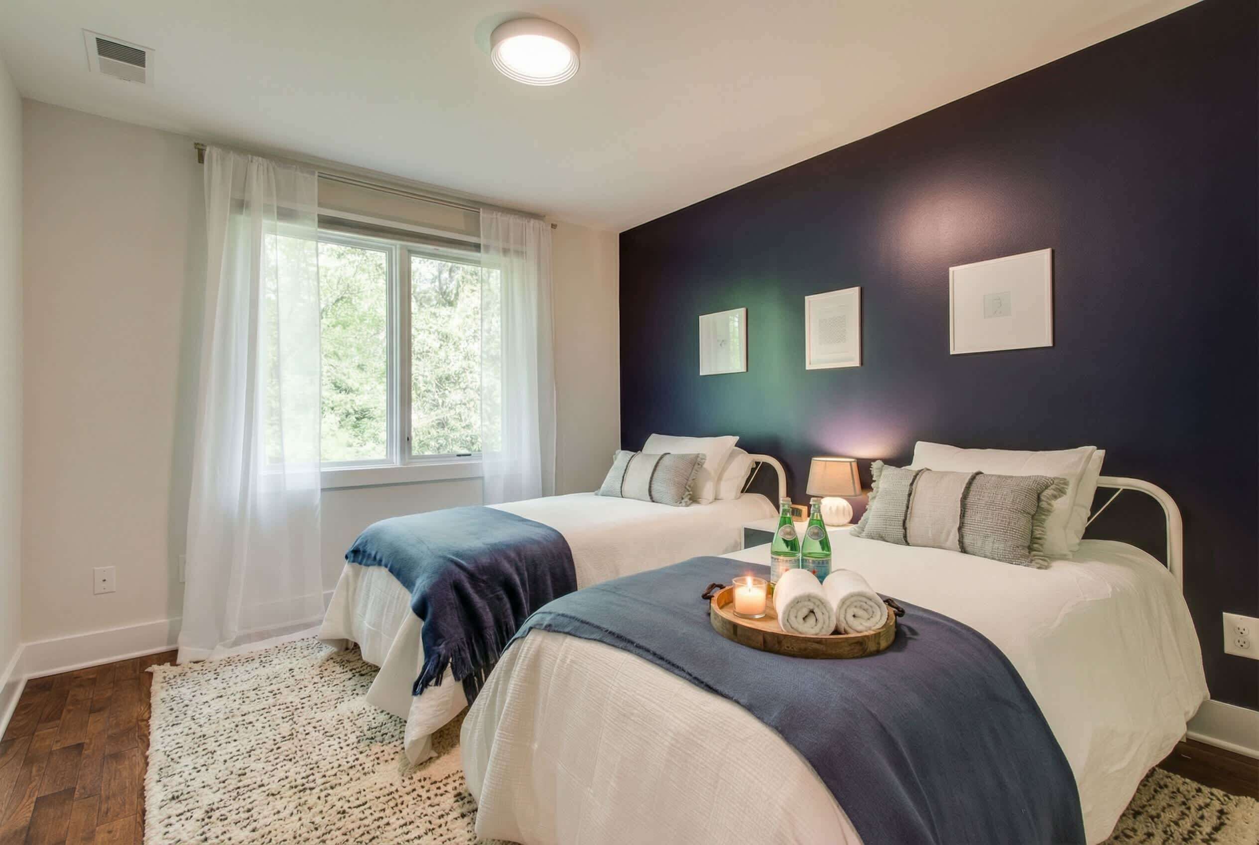 Quarto com duas camas, uma janela grande com cortinas leves e uma parede escura com quadros.