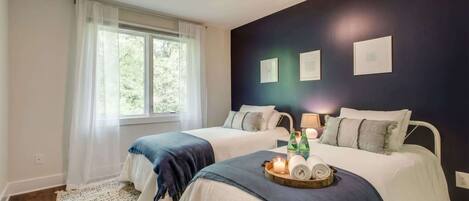Quarto com duas camas, uma janela grande com cortinas leves e uma parede escura com quadros.