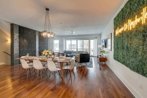 A modern dining area with a wooden table set for six, a black sofa, and a green wall with a neon sign.
