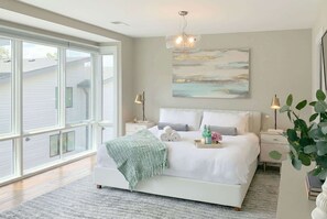 Un dormitorio con un ventanal grande, una cama con ropa de cama blanca, dos mesitas de noche y una obra de arte colgada en la pared.