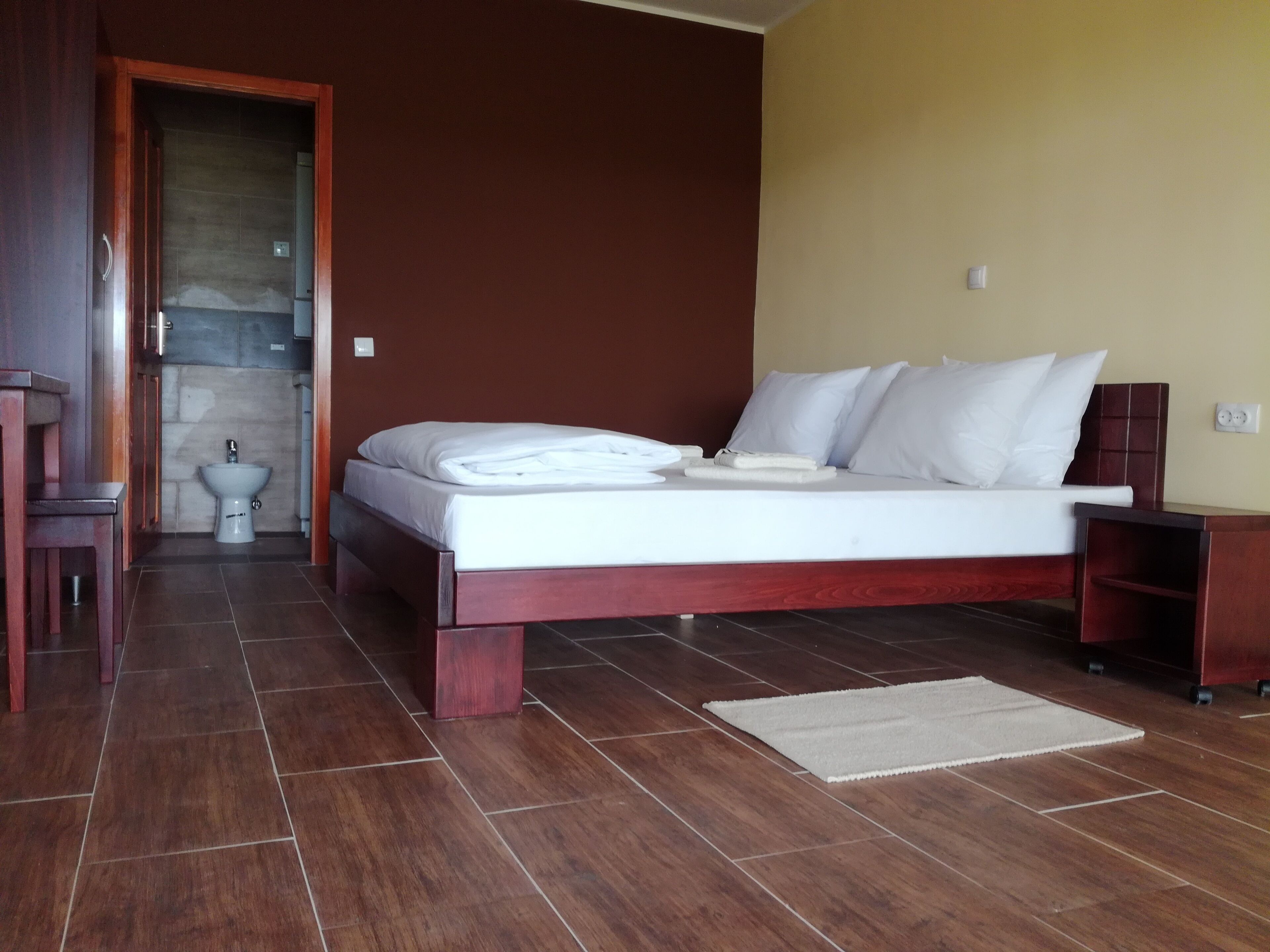 A bedroom with a bed, a wooden nightstand, a bedside table, a chair, and a bathroom with a toilet and sink.
