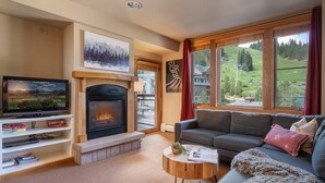 A living room with a fireplace, a television, a sofa, and large windows with a view of a green landscape. - A+ Base Area Condo | Zephyr Mountain Lodge | Mountain Views | New Decor | Ski In Ski Out (Winter Park)