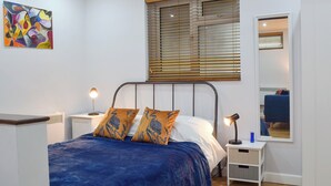 Een slaapkamer met een bed, twee nachtkastjes, een wandspiegel en een kleurrijk abstract schilderij. Er is ook een raam met zonwering.