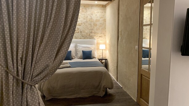 A bedroom with a bed, a nightstand, a lamp, and a view of a stone wall and a wooden door. - Le Petit Hameau de Chantilly - Chic and Charming Studio in the Heart of Chantilly (CHANTILLY)