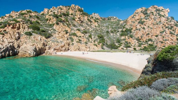 A coastal beach with clear turquoise waters, rocky cliffs, and a sandy shore.