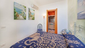 A bedroom with a bed, two paintings, an air conditioner, a chair, and a view of a kitchen.