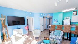 Una sala de estar con televisión de pantalla plana, muebles de mimbre, un armario blanco y un refrigerador.