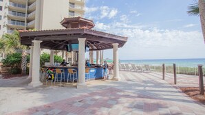 Zona de bar al aire libre techada con vista a la piscina, barra y área de estar con sillas.