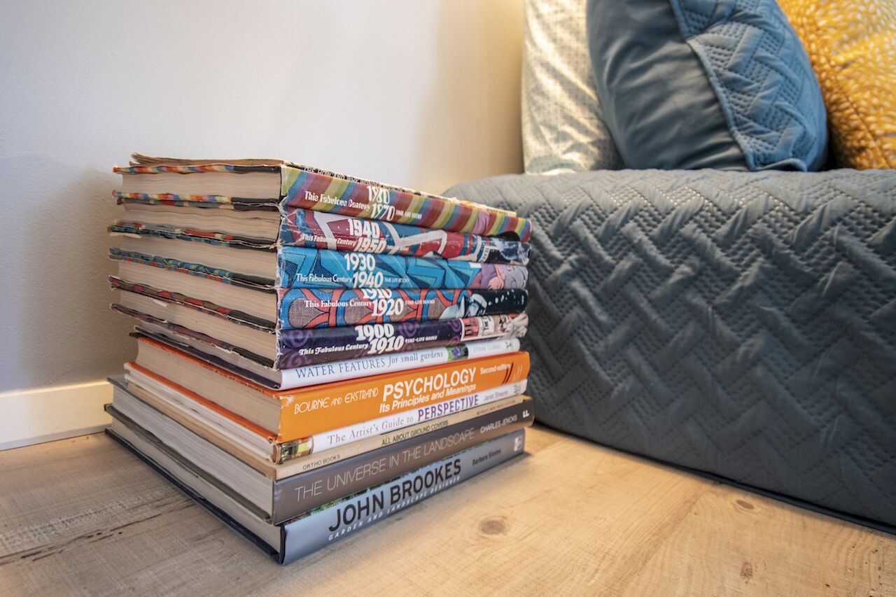 A stack of books on a wooden floor next to a grey couch with a blue pillow.