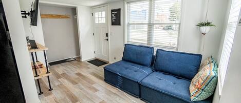 Een kamer met een blauwe bank, een kleine tafel met een laptop, een aan de muur bevestigde televisie en een raam met zonwering.