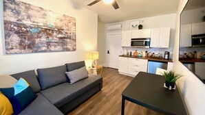 Un apartamento moderno con un sofá gris, una mesa de comedor, un ventilador de techo y una cocina con armarios y electrodomésticos blancos.