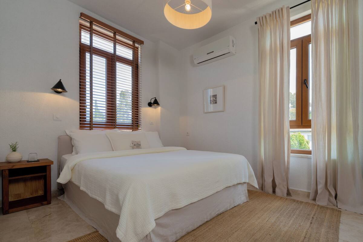 A bedroom with a bed, a nightstand, a wall-mounted air conditioner, and a window with blinds.