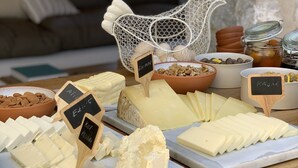 A cheese board with various types of cheese and labels, set against a stone wall and a couch in the background. - The River House (Ula)