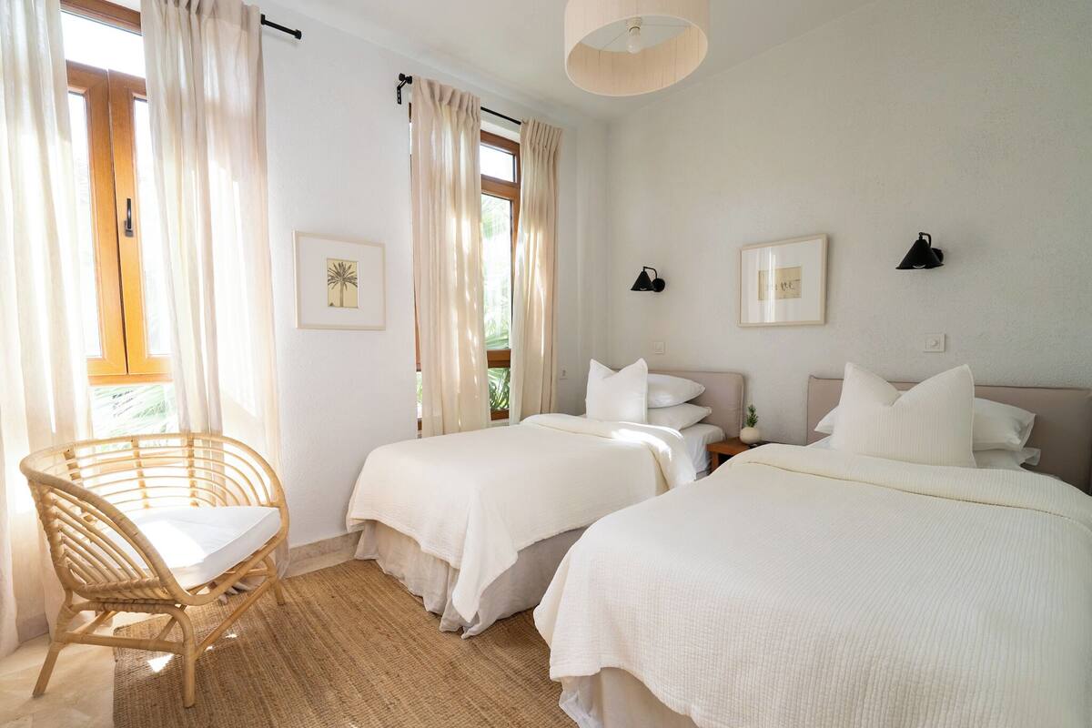 A bedroom with two beds, a wicker chair, and a small table.