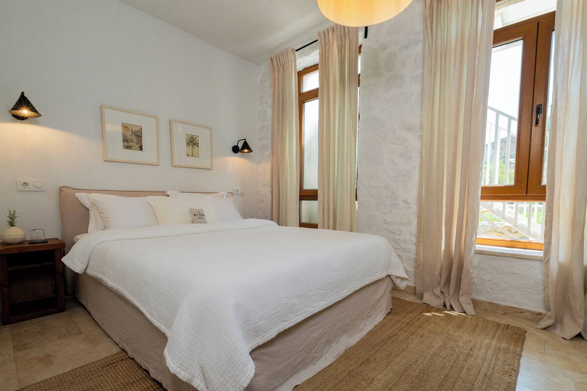 A bedroom with a bed, bedside tables, a lamp, a window with curtains, and framed pictures on the wall.