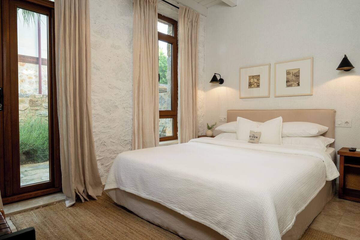 A bedroom with a large bed, beige curtains, a small bedside table, and framed pictures on the wall.