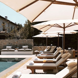 A poolside area with lounge chairs and umbrellas, a stone wall, and a building in the background. - The River House (Ula)